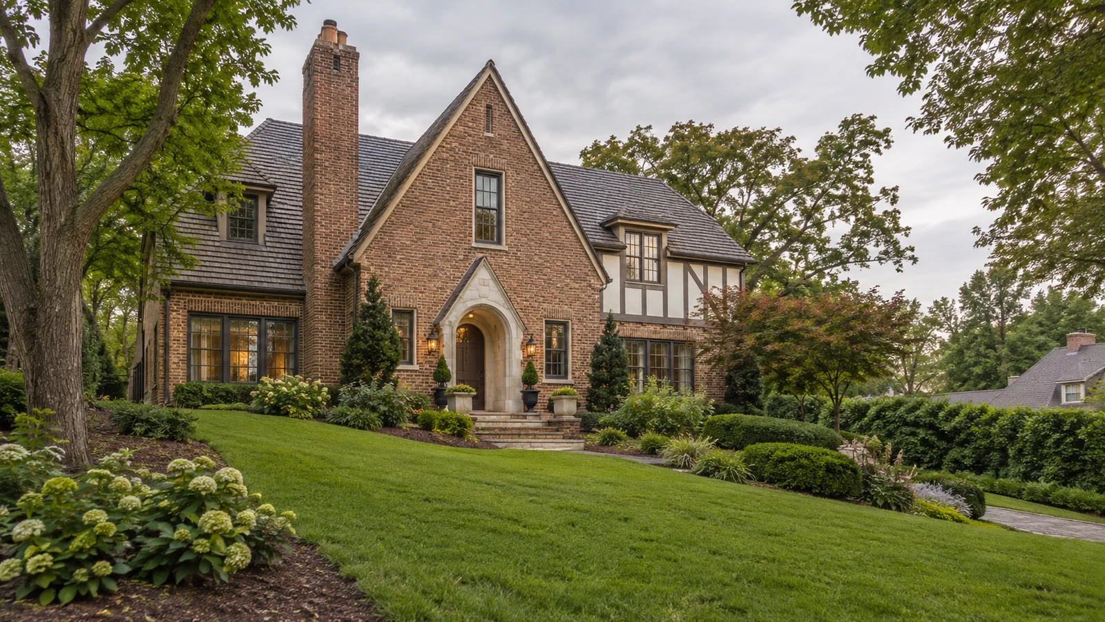 An Alabama Tudor-inspired home with warm brick, a sloping landscaped garden, property boundary hedge and neighboring residential roof in the distance, elegant overcast light. Context for researching property and county costs.